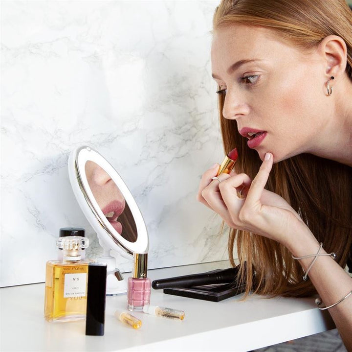 A woman with long red hair applies lipstick, focusing on her reflection in the UNIQ PRO BEAUTY Bathroom Mirror with LED Light and Suction Cup x10 Magnifying Mirror (White) on a white table with perfume, nail polish, and makeup items.